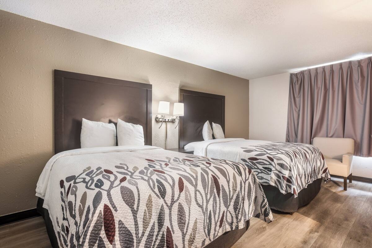 A hotel room with two beds, a nightstand with lamps, a chair, and a large window with curtains.