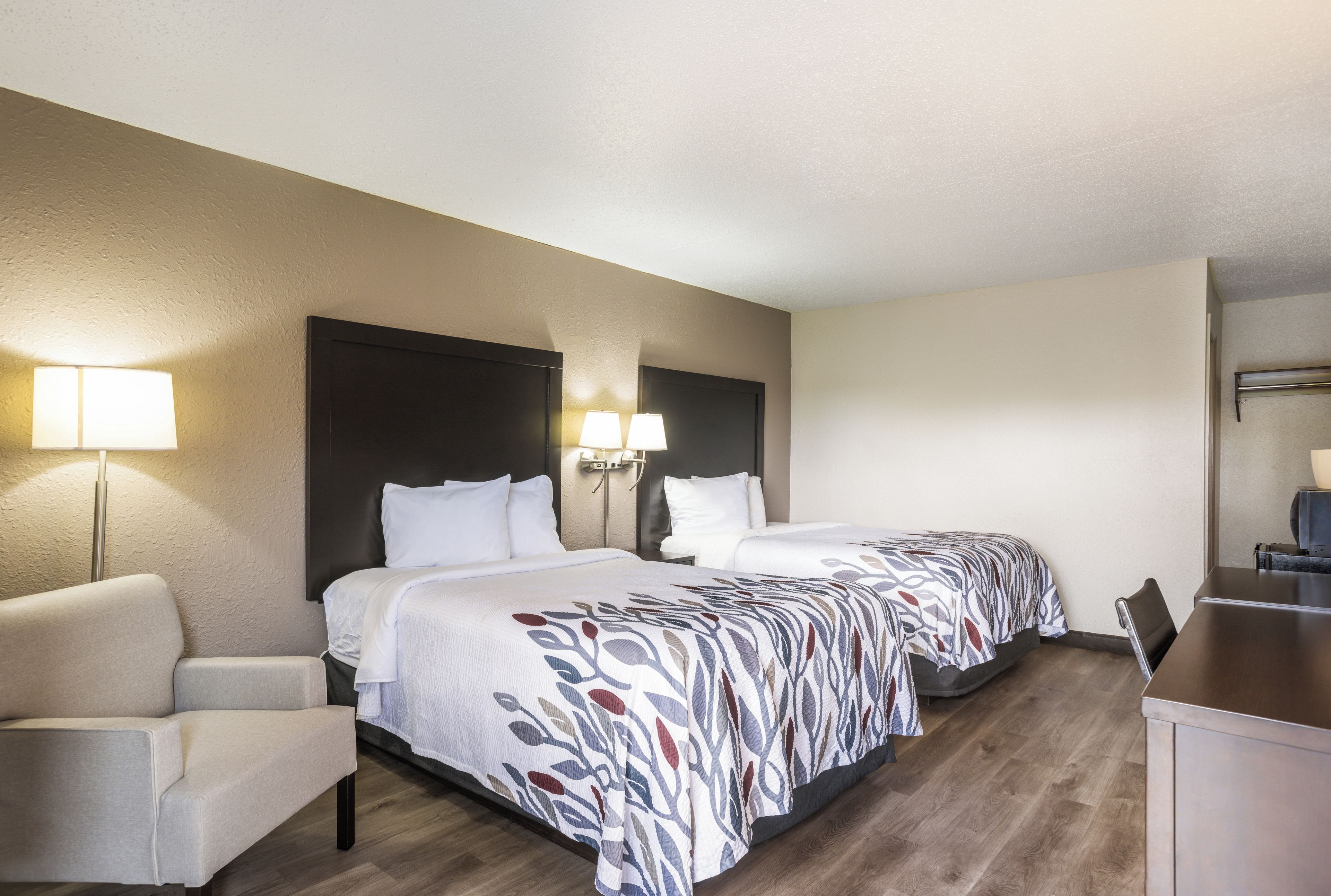 A hotel room with two beds, a desk, a chair, and a lamp.