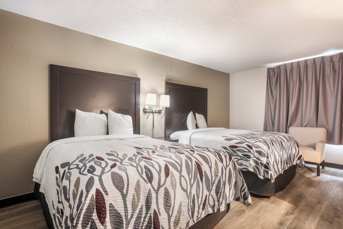 A hotel room with two beds, a nightstand with lamps, a chair, and a large window with curtains.