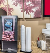 Máquina de café com ecrã digital, copos de papel empilhados e dispensador de café sobre um balcão.