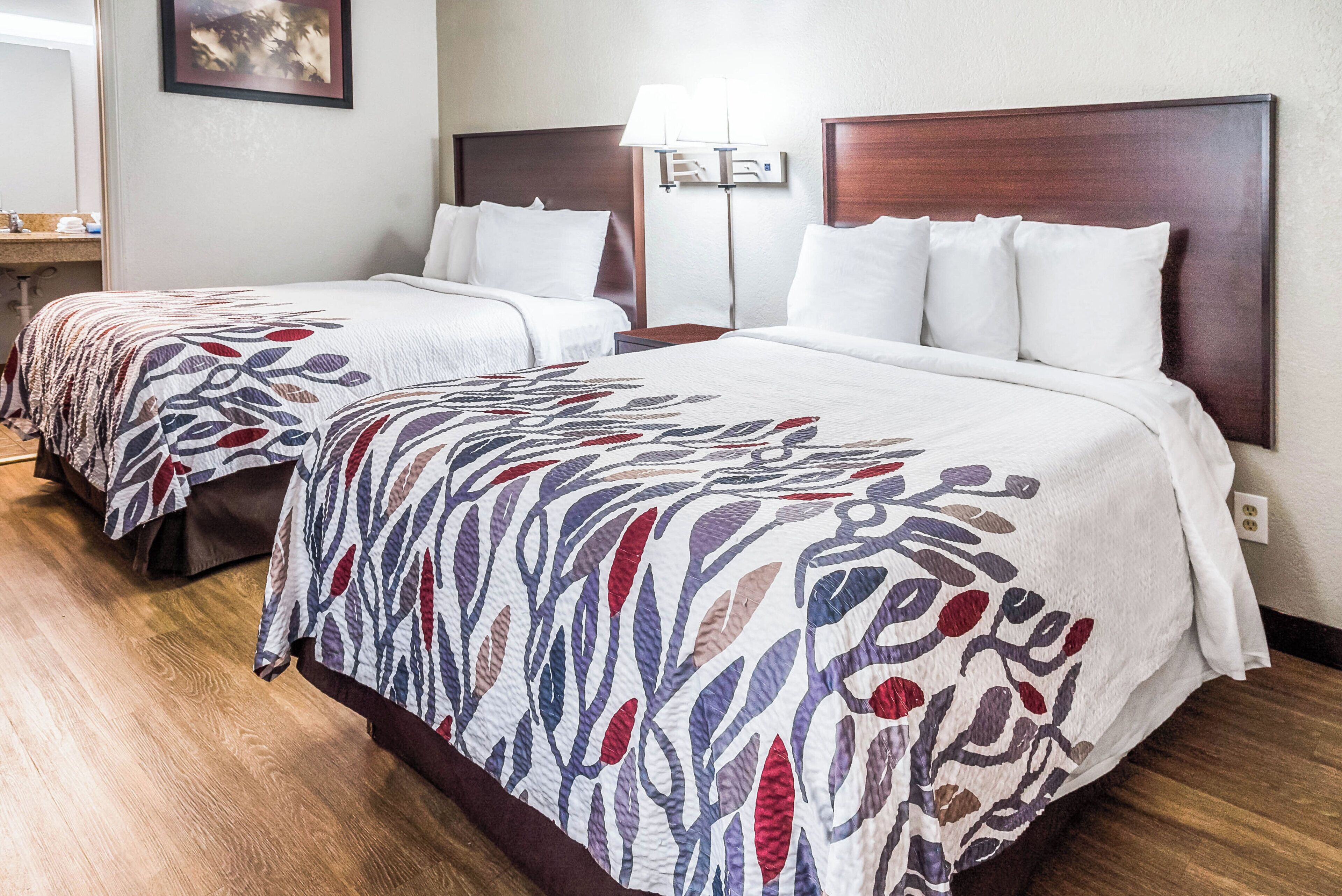 Twin beds with patterned bedspreads, a bedside table, a lamp, and a wall-mounted light fixture in a hotel room.