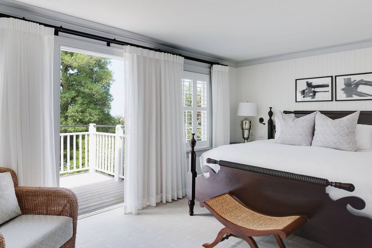 A bedroom with a large bed, a wooden chair, a balcony with white curtains, and a view of greenery.