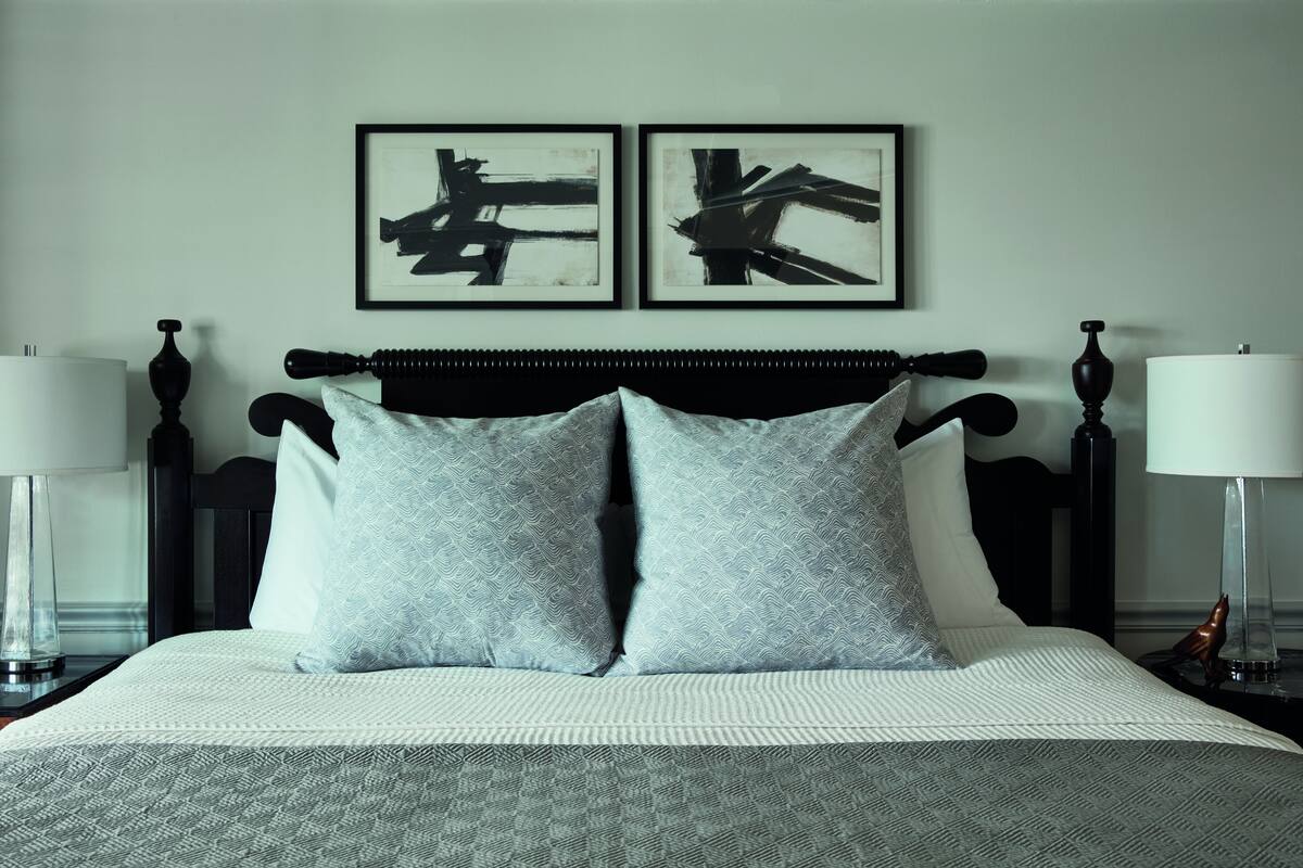 A neatly made bed with a patterned bedspread and two pillows, flanked by nightstands with lamps and a decorative item. Two framed abstract paintings hang above the bed.