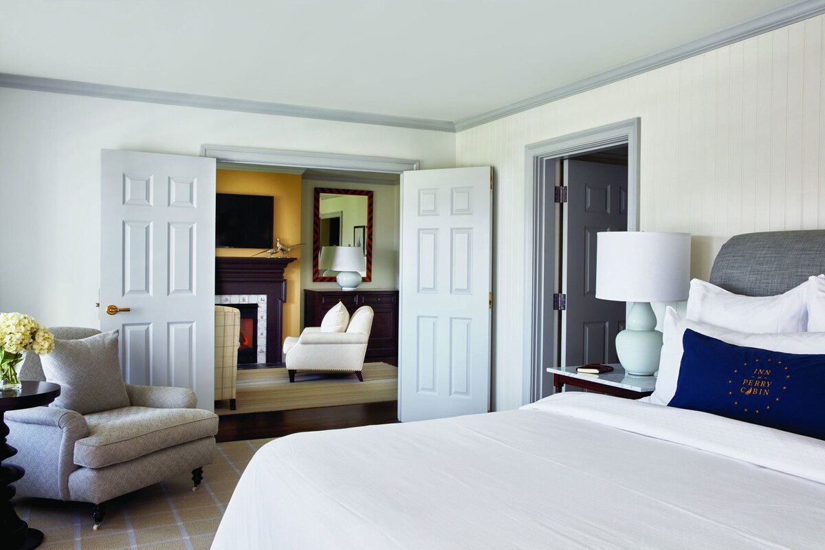 A hotel room with a bed, a chair, a desk, a lamp, and a fireplace.