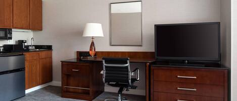 A hotel room with a kitchenette, a desk with a chair, a flat-screen TV, and a large mirror on the wall.