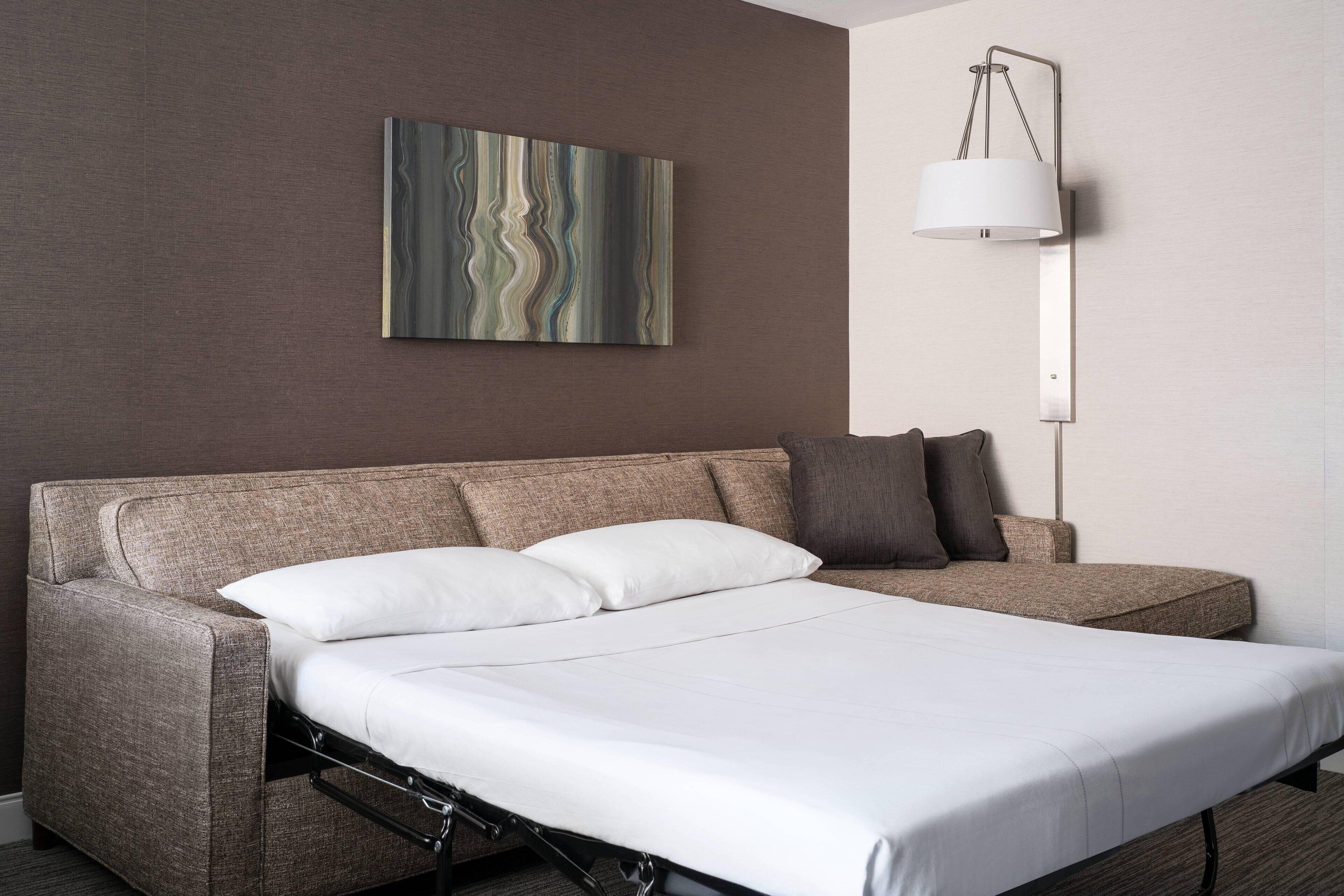 A modern hotel room with a sofa bed, a wall-mounted lamp, and a piece of abstract art.