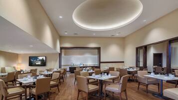 A hotel dining area with multiple tables and chairs, a large screen, and a modern ceiling design.