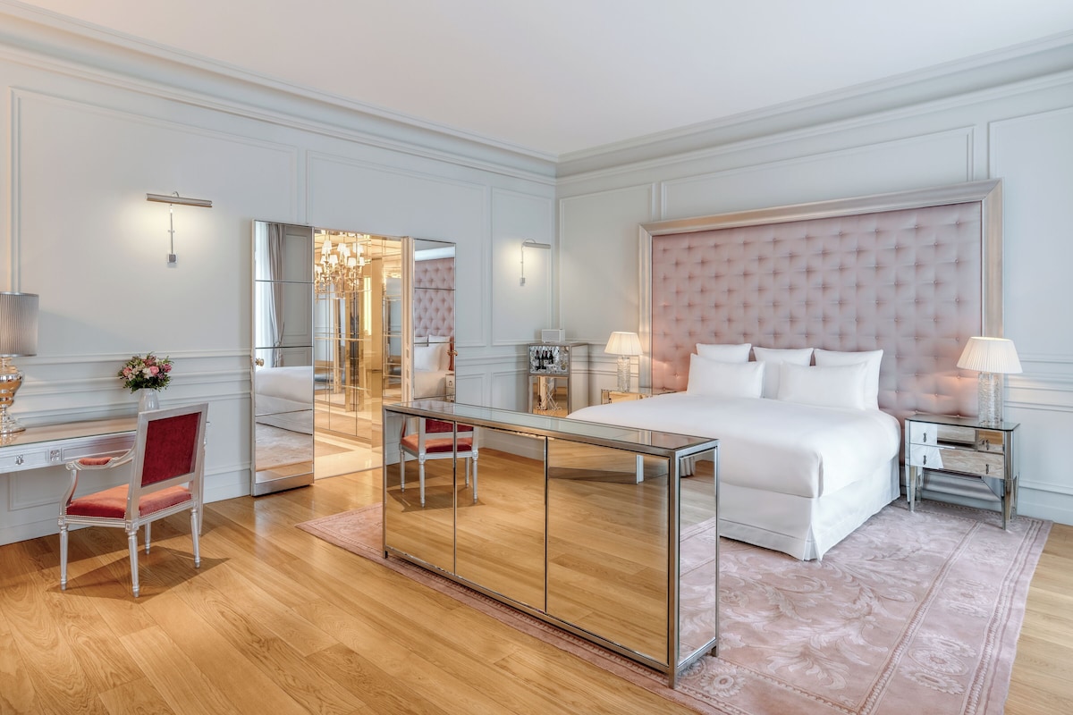 A spacious bedroom with a large bed, a sitting area with a chair, a desk, and a mirror.