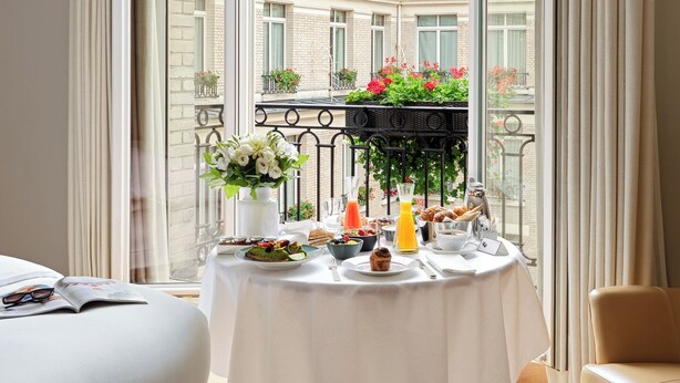 Un balcón con una mesa dispuesta para el desayuno, con vista a una zona residencial con balcones y jardineras.