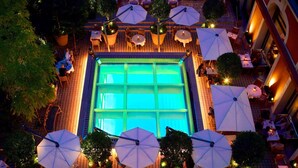 Una piscina al aire libre con mesas y sombrillas alrededor, rodeada de plantas en macetas y árboles.