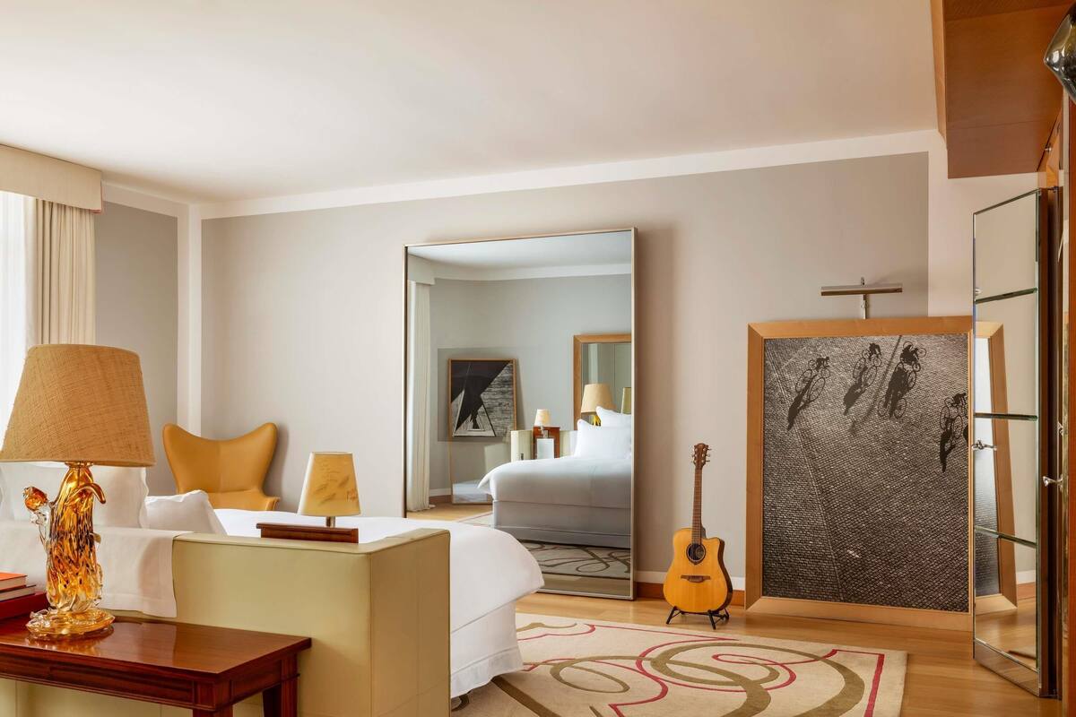 A hotel room with a bed, a sitting area with a table and chairs, a guitar, a large mirror, and artwork on the walls.