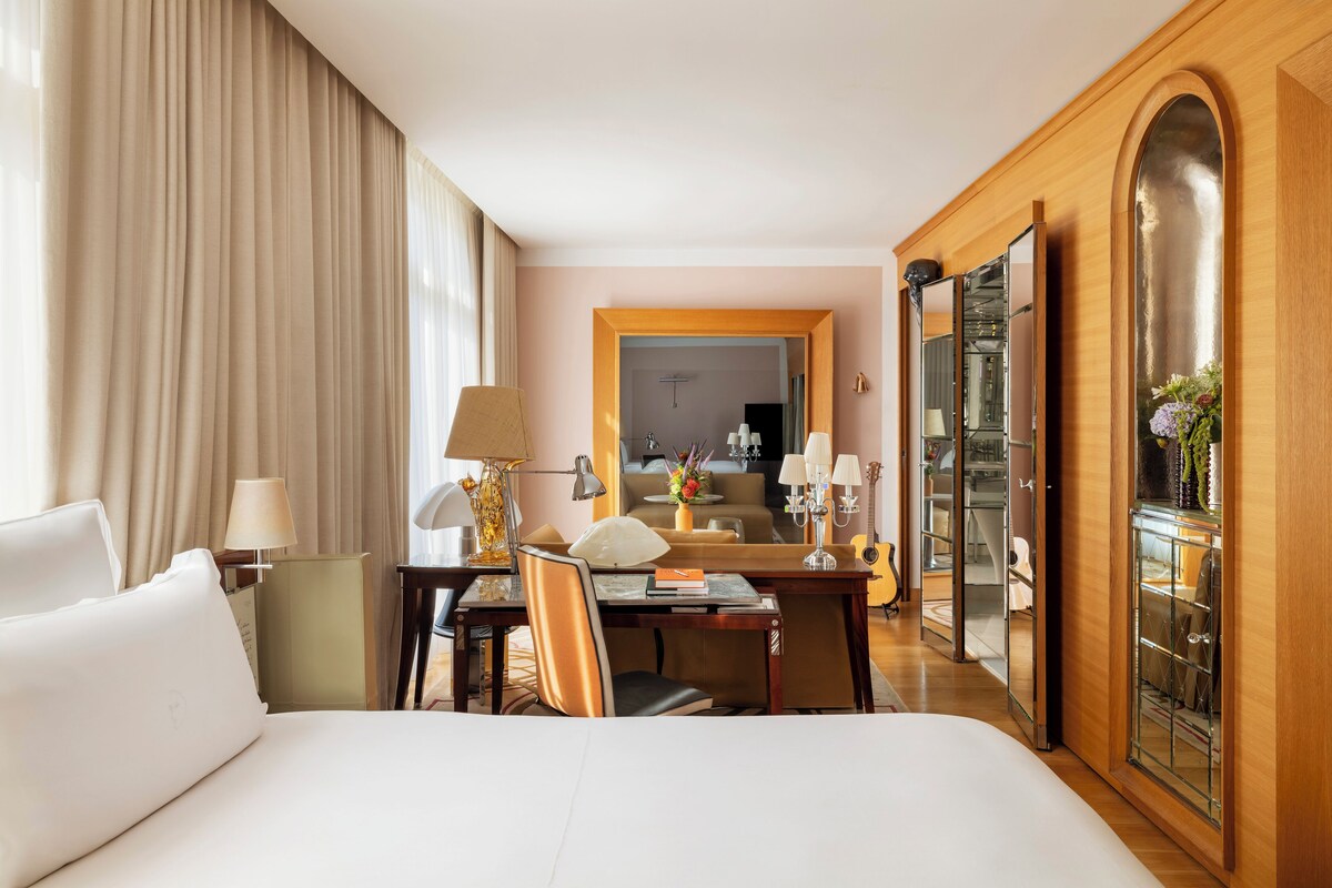 A hotel room with a large bed, a desk with a lamp, a chair, a mirror, and a wardrobe.