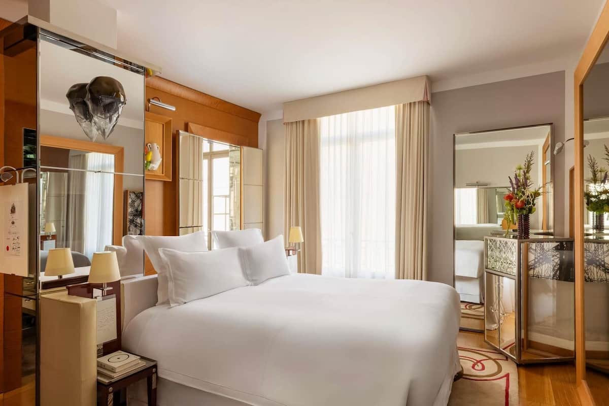 Habitación de hotel con una cama grande, una mesita de noche con lámparas, un espejo y vistas a la ciudad.