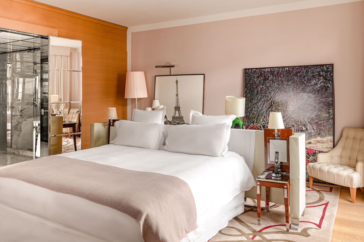 A hotel room with a large bed, a desk with a lamp, a chair, a glass shower enclosure, and artwork on the walls.
