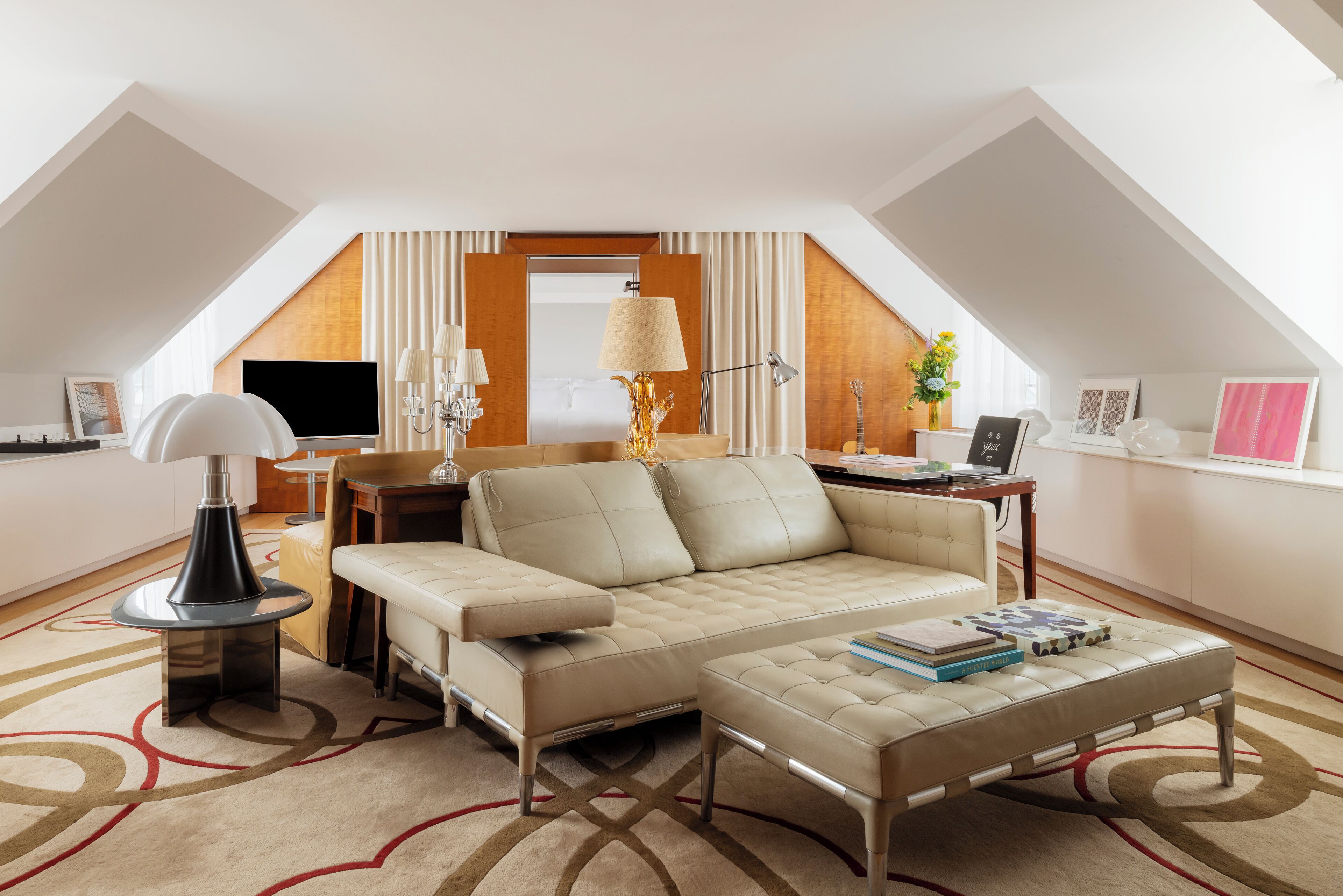 A modern hotel room with a loft-style ceiling, a sofa, an ottoman, a desk, and a television.