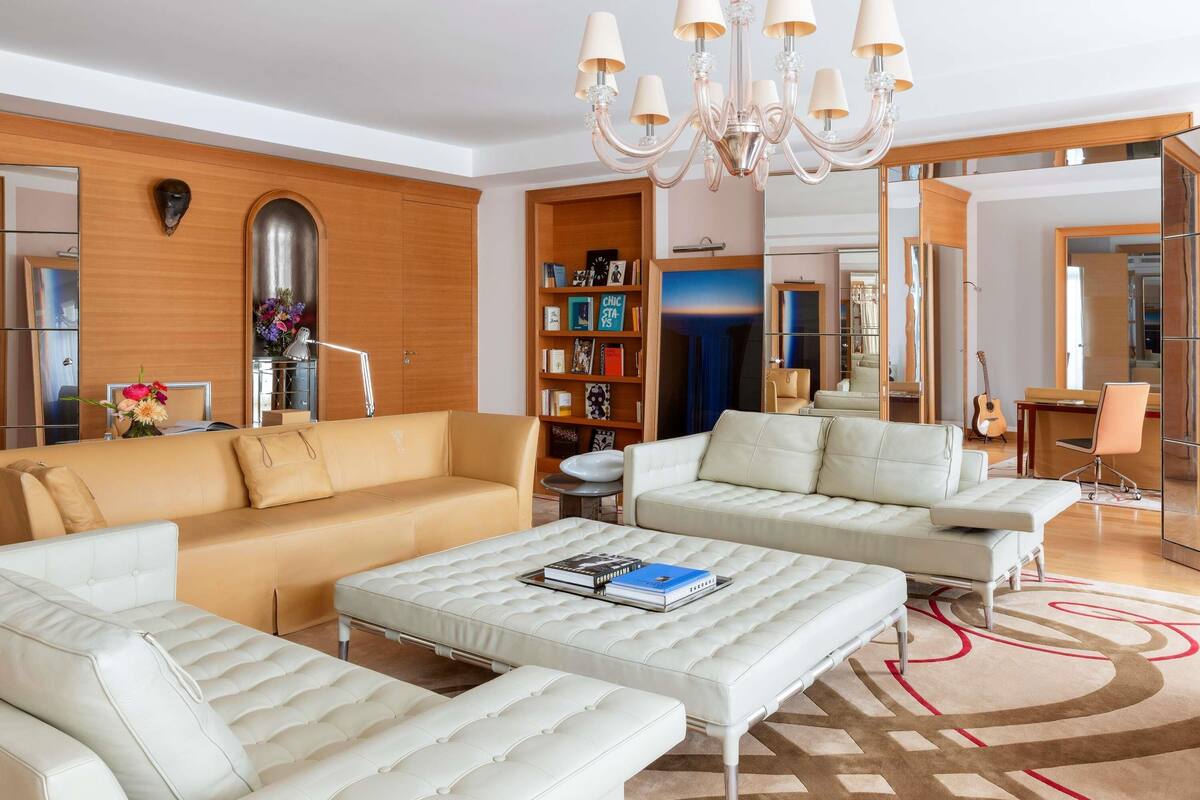 A modern living room with a beige sofa, a white sofa, a white ottoman, a chandelier, a bookshelf, and a mirror.