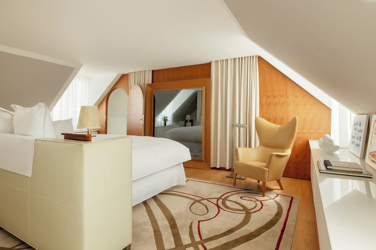 A modern hotel room with a large bed, a sitting area with a chair, a desk, and a view of the city through a large window.