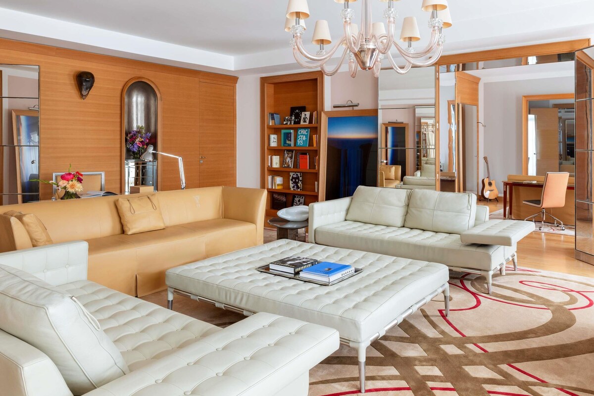 Una sala de estar moderna con un sofá color beige, un sofá blanco, un otoman blanco, una araña, una estantería y un espejo.