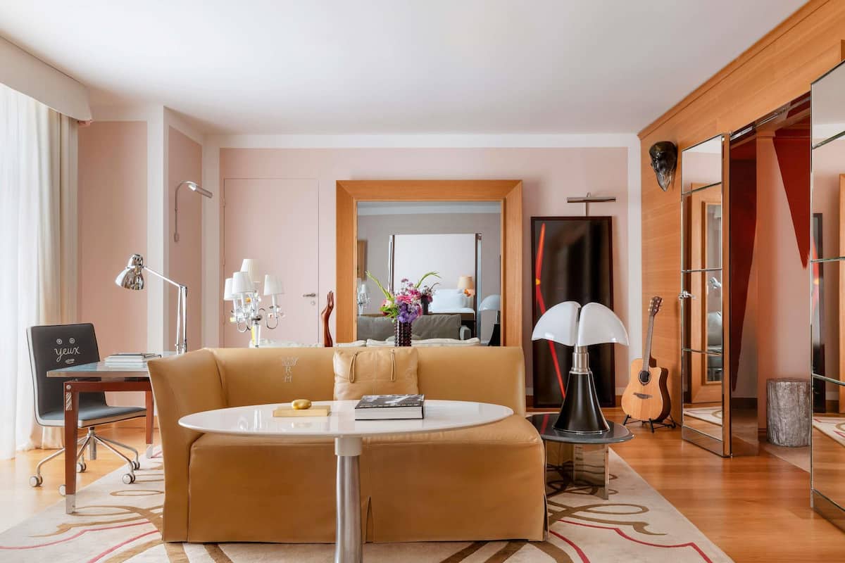 A modern hotel room with a sofa, a round table, a chair, a lamp, a guitar, and a mirror.