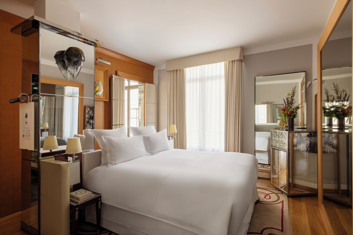 A hotel room with a large bed, a desk with a lamp, a mirror, and a view of the city through large windows.