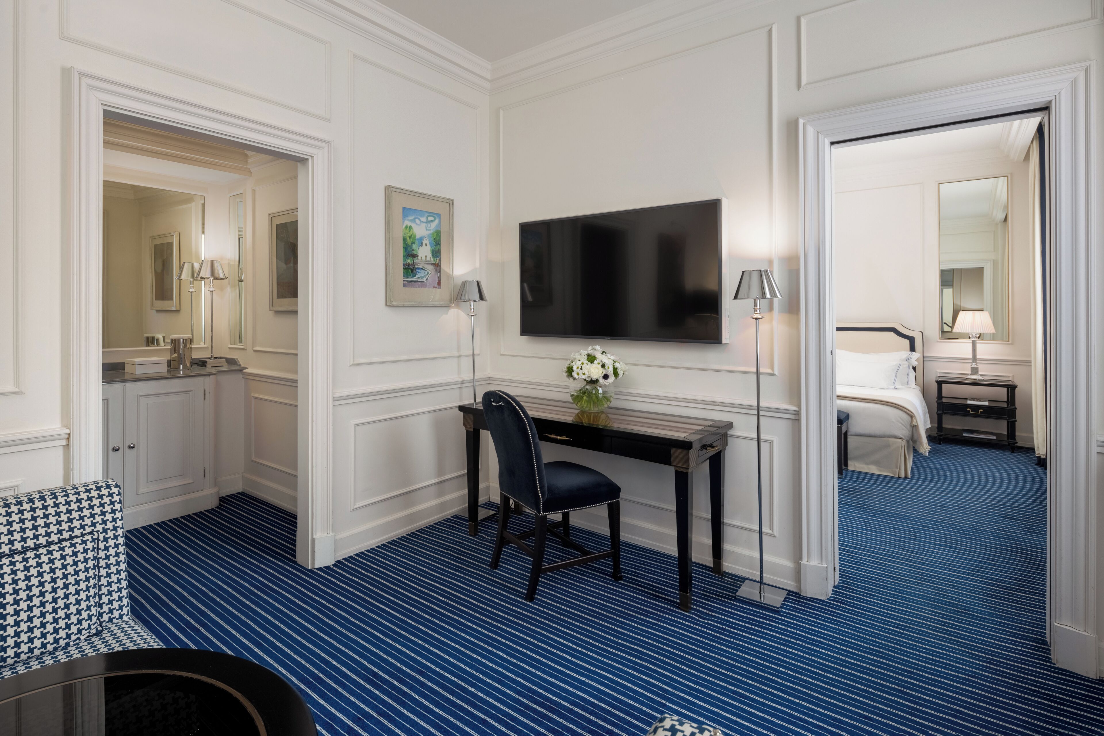 A hotel room with a blue-striped carpet, a desk with a chair, a flat-screen TV, a bed, and a small table with a lamp.