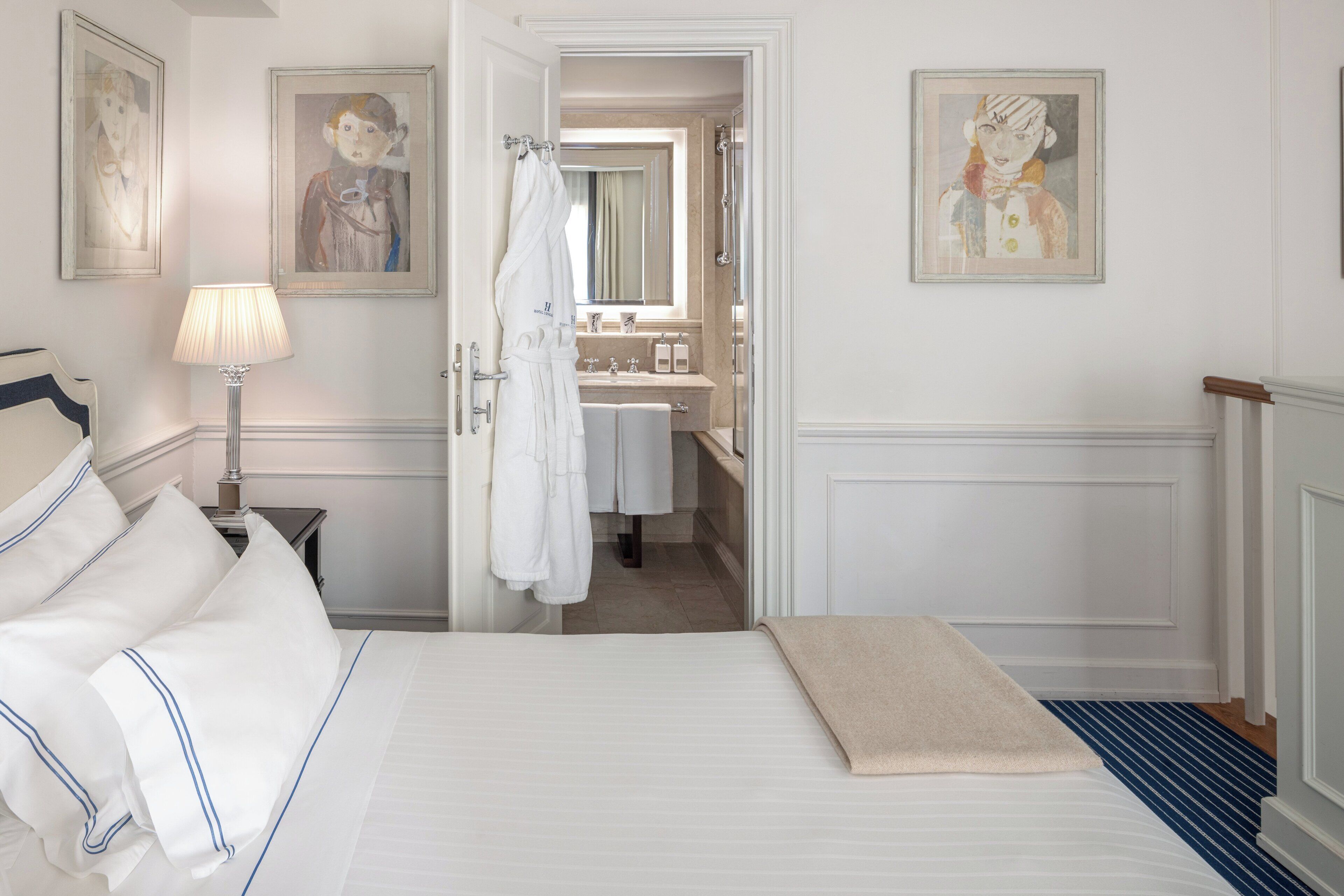 Ein Hotelzimmer mit einem ordentlich bezogenen Bett, einer Nachttischlampe, einem Badezimmer mit Waschbecken und Spiegel sowie Bildern an den Wänden.