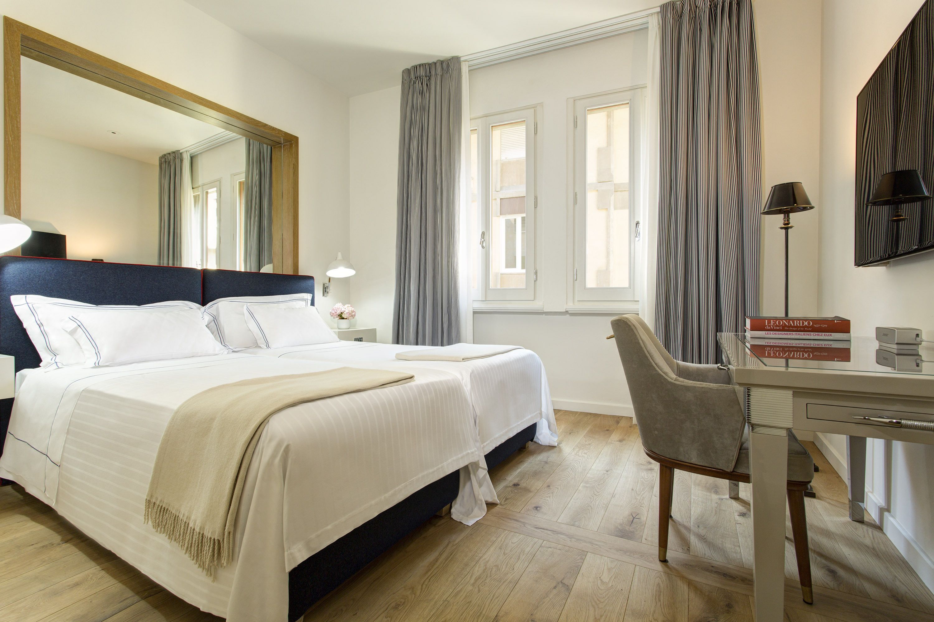 Habitación de hotel con una cama grande, un escritorio y una silla. Hay un espejo en la pared, una lámpara en el escritorio y cortinas en las ventanas.