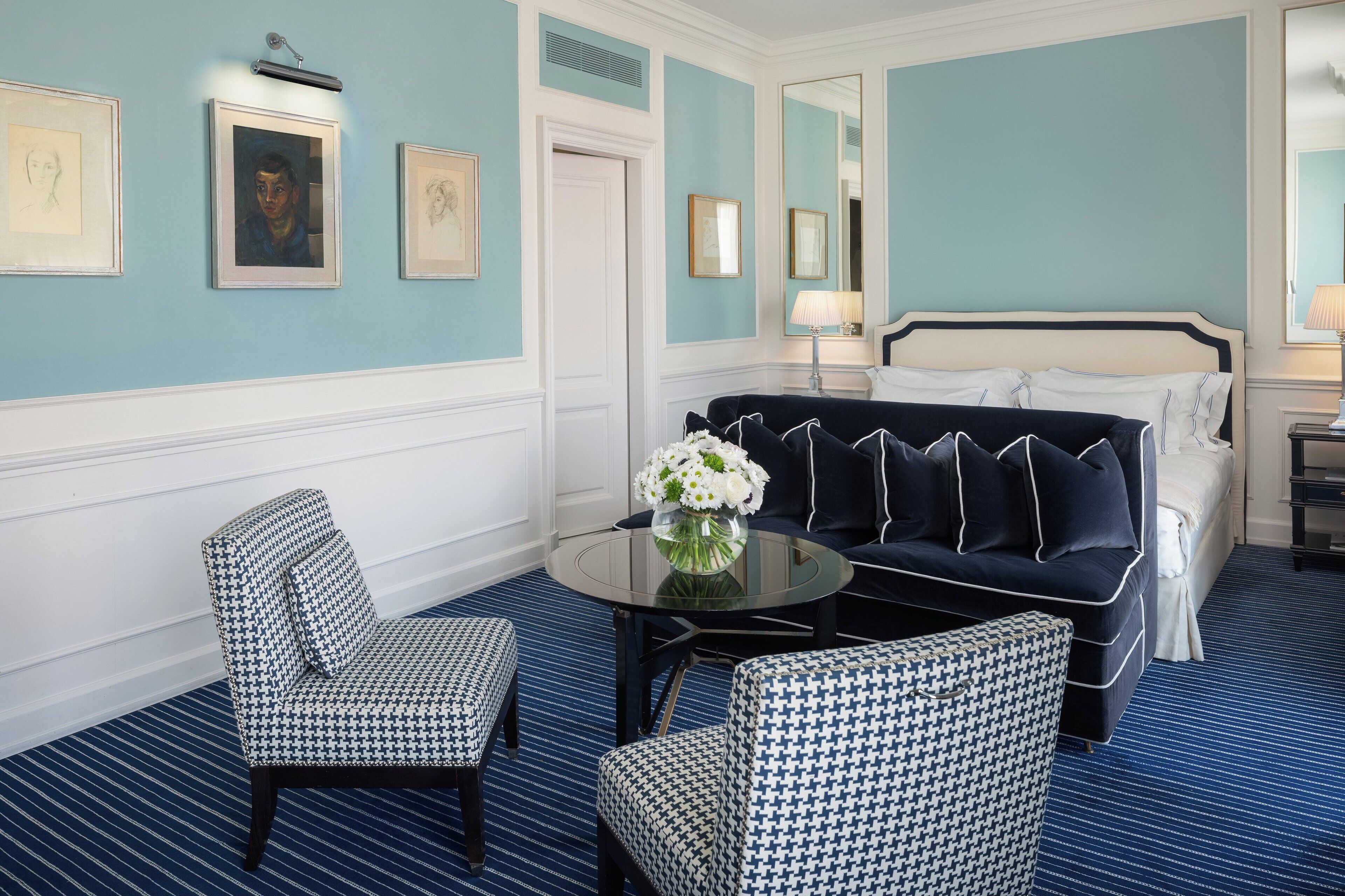 A hotel room with a blue and white color scheme, featuring a bed, a sofa, two patterned armchairs, a round table with a vase of flowers, and framed artwork on the walls.