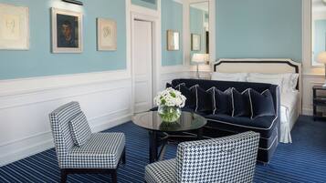 A hotel room with a blue and white color scheme, featuring a bed, a sofa, two patterned armchairs, a round table with a vase of flowers, and framed artwork on the walls.