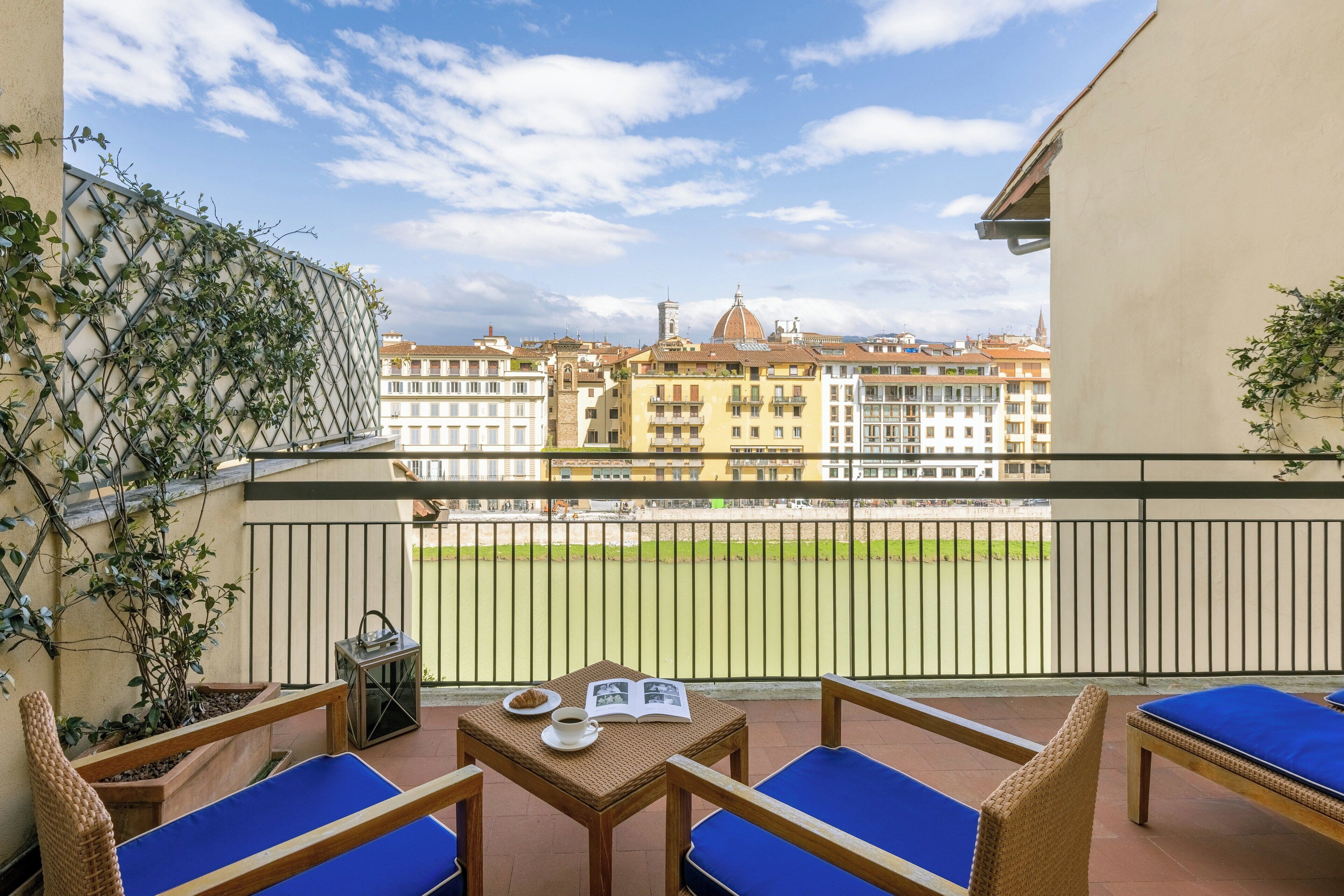 A balcony with blue cushioned chairs, a small table with a book and a cup, and a view of a sports field and buildings in the background.