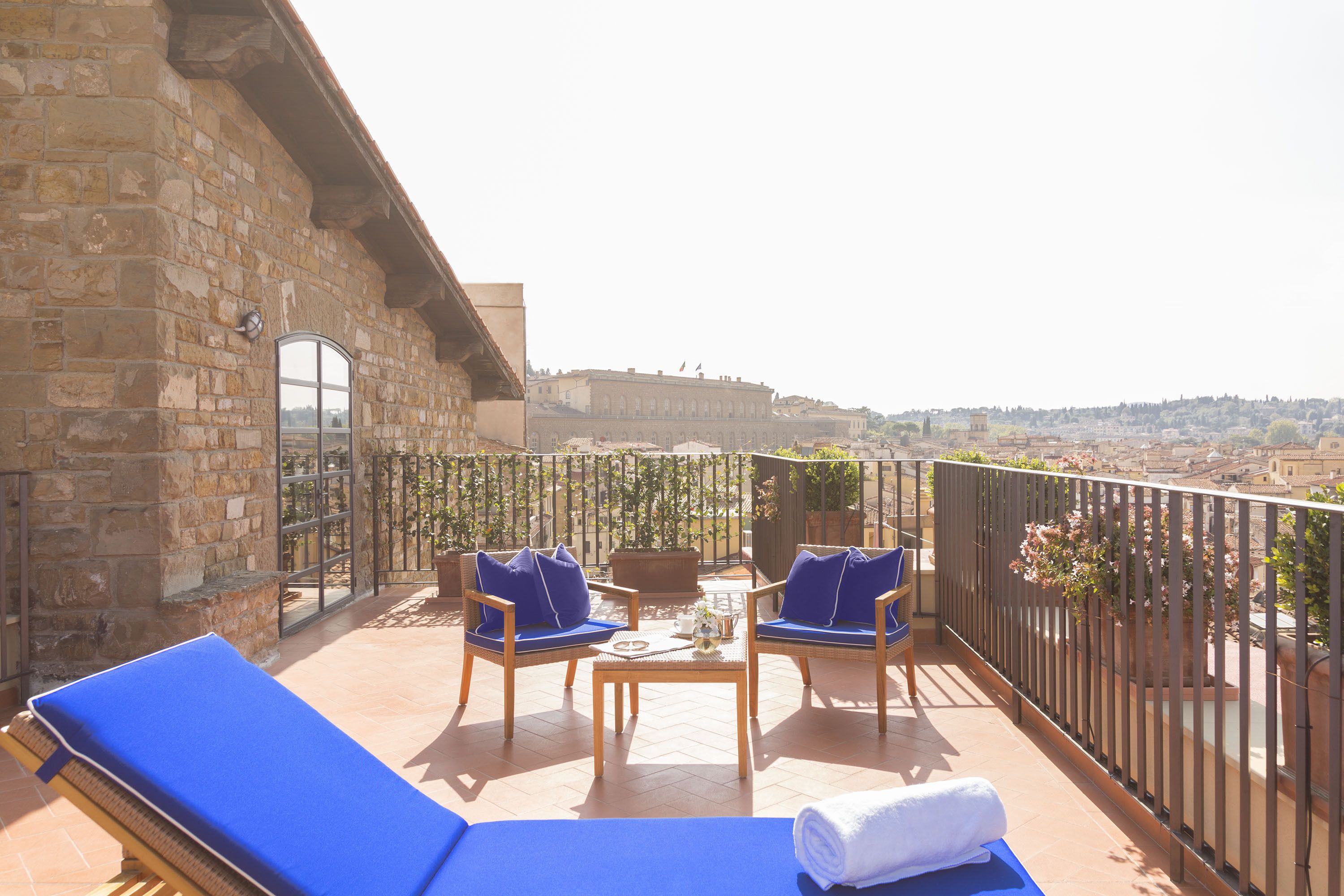 A balcony with two blue lounge chairs, a small table, and a view of a historic building in the distance.
