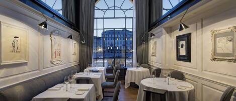 Un restaurante con varias mesas, vista a un edificio a través de grandes ventanales y cuadros en las paredes.