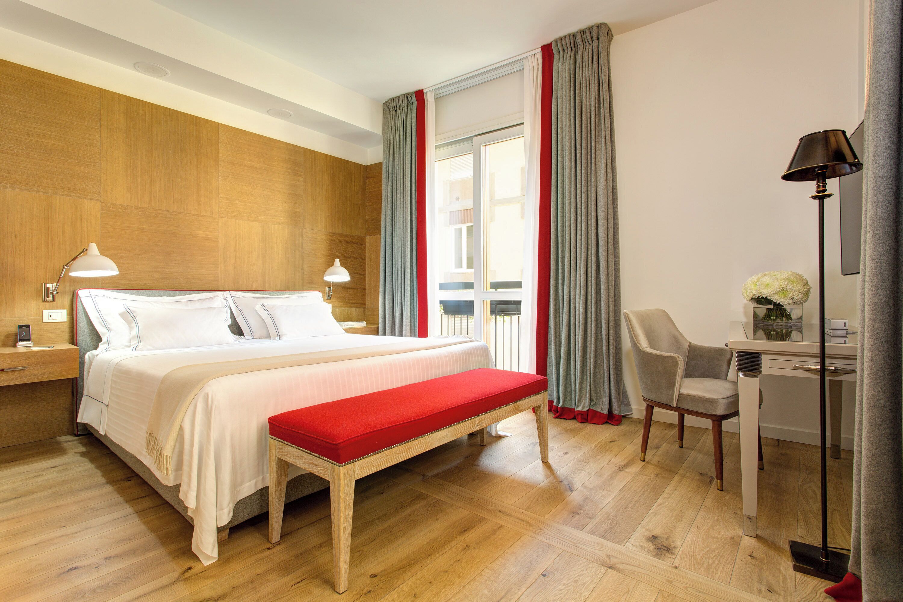 A hotel room with a wooden floor, a bed with white bedding, a red bench, a desk with a chair, and a lamp.