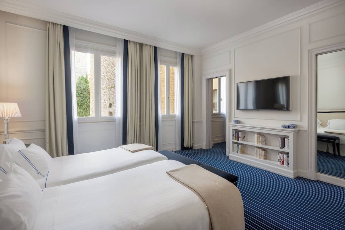 Hotellihuone, jossa on kaksi sänkyä, televisio, kirjahylly ja suuri ikkuna, jossa on verhot.