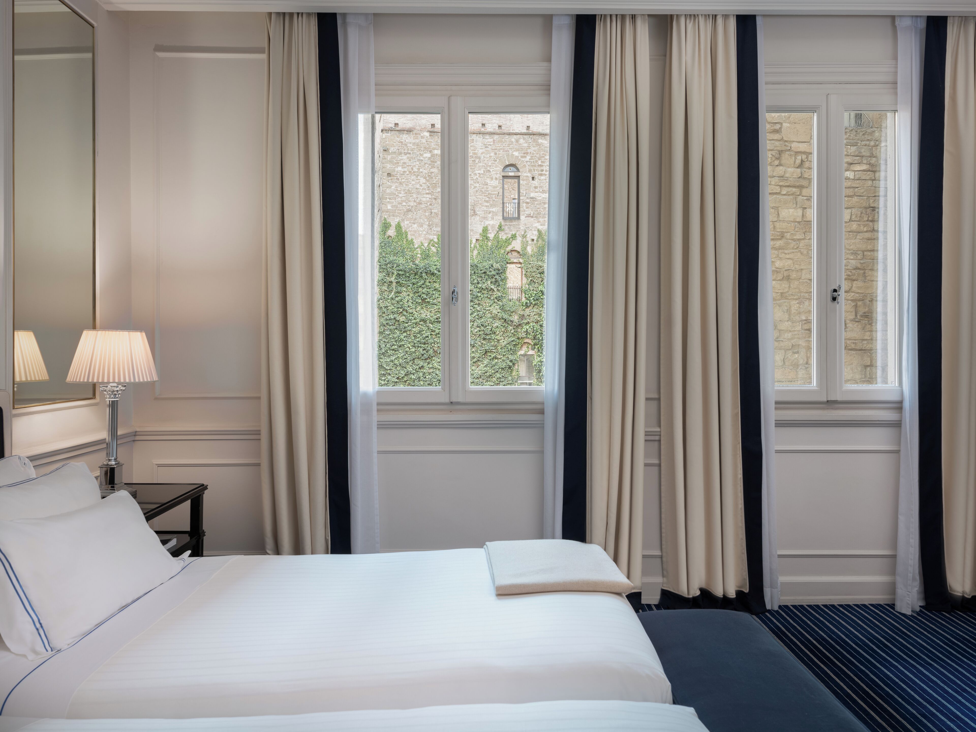 A hotel room with a large bed, a bedside table with a lamp, two windows with curtains, and a view of greenery and a stone building outside.