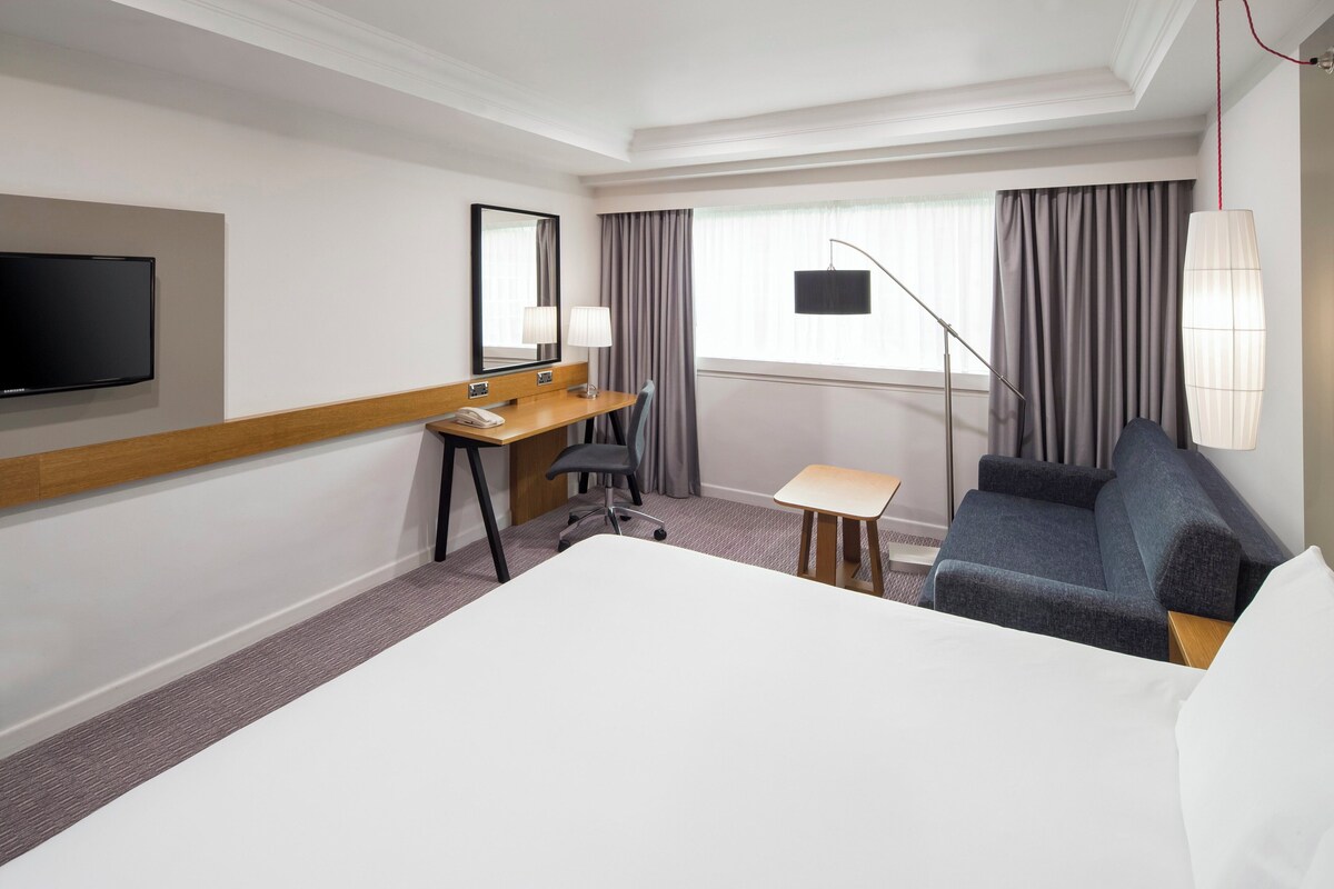 Ein Hotelzimmer mit einem Bett, einem Schreibtisch, einem Stuhl, einem Fernseher und einem fensterbepflanzten Fenster.