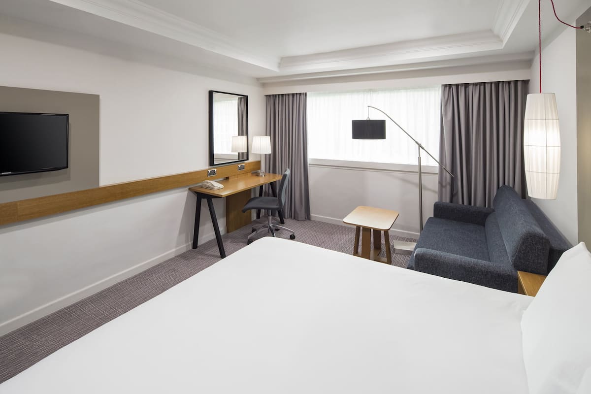A hotel room with a large bed, a desk with a telephone, a chair, a sofa, a TV mounted on the wall, and a window with curtains.