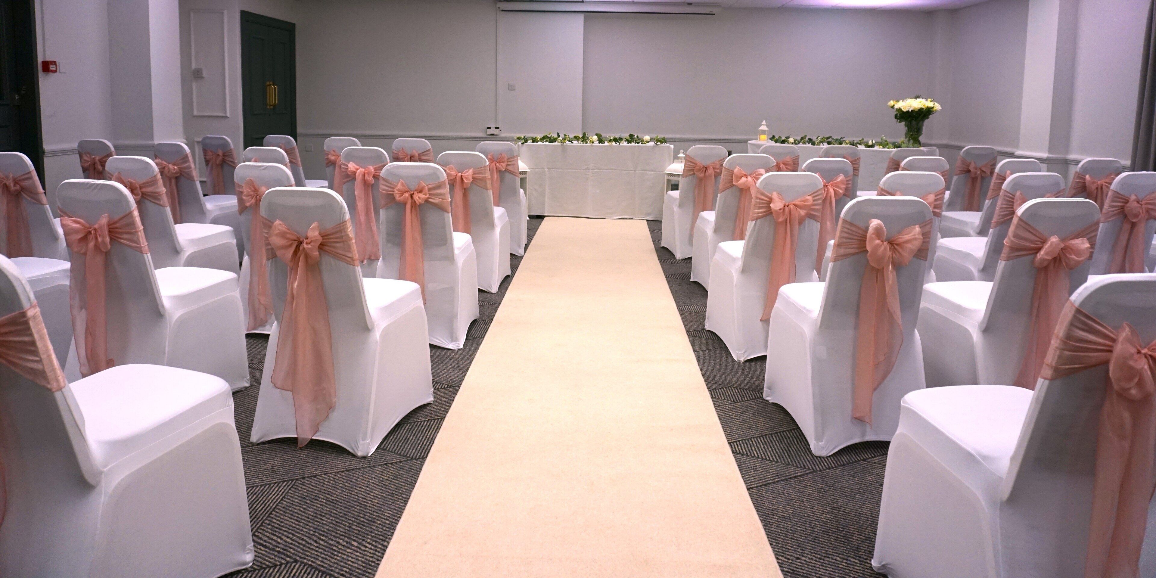 Un salón de conferencias con filas de sillas blancas y lazos color melocotón, un pasillo alfombrado en tonos beige y una mesa blanca al frente.