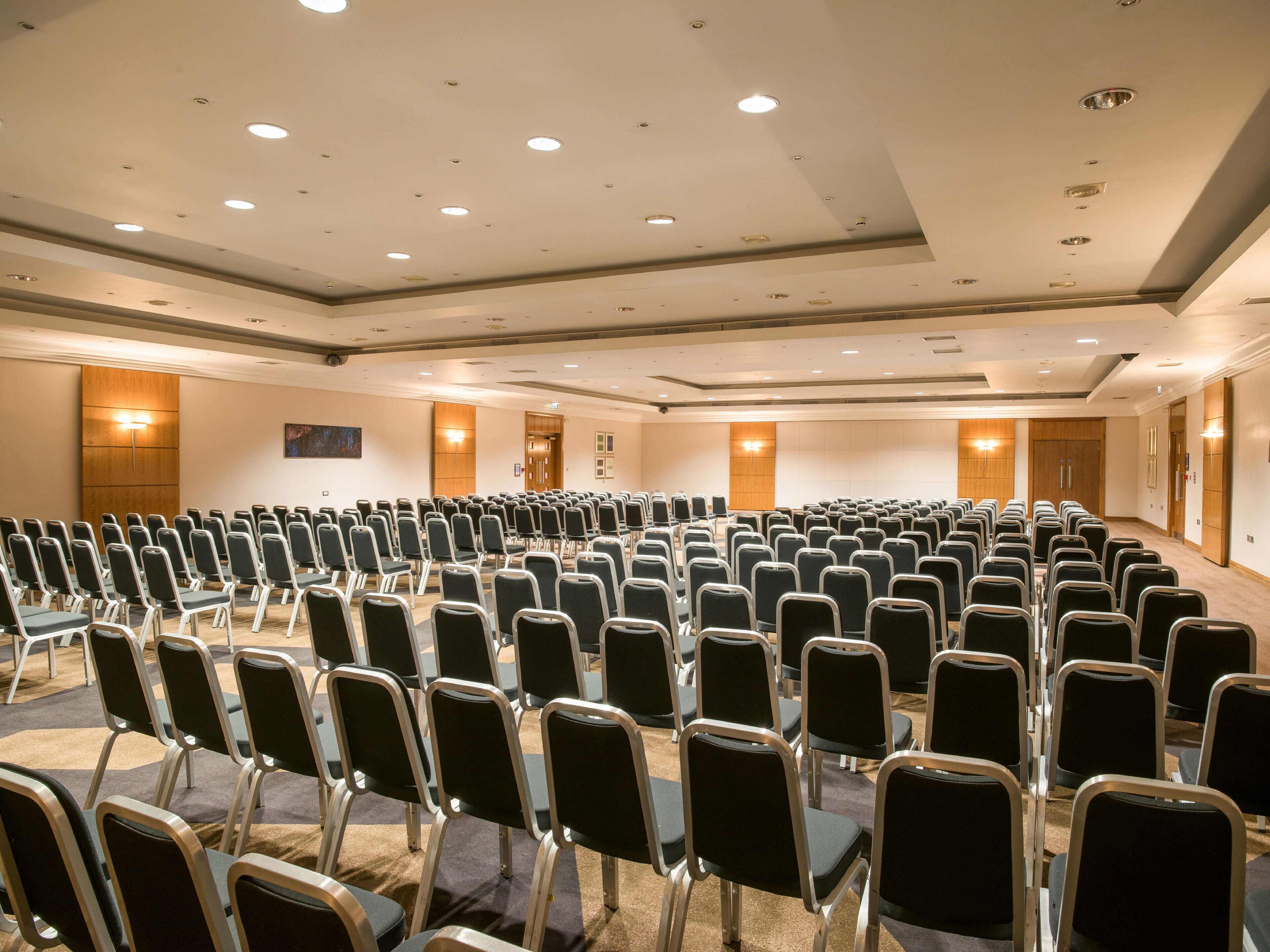 A conference room with rows of chairs, a wooden panel wall, and recessed ceiling lights.