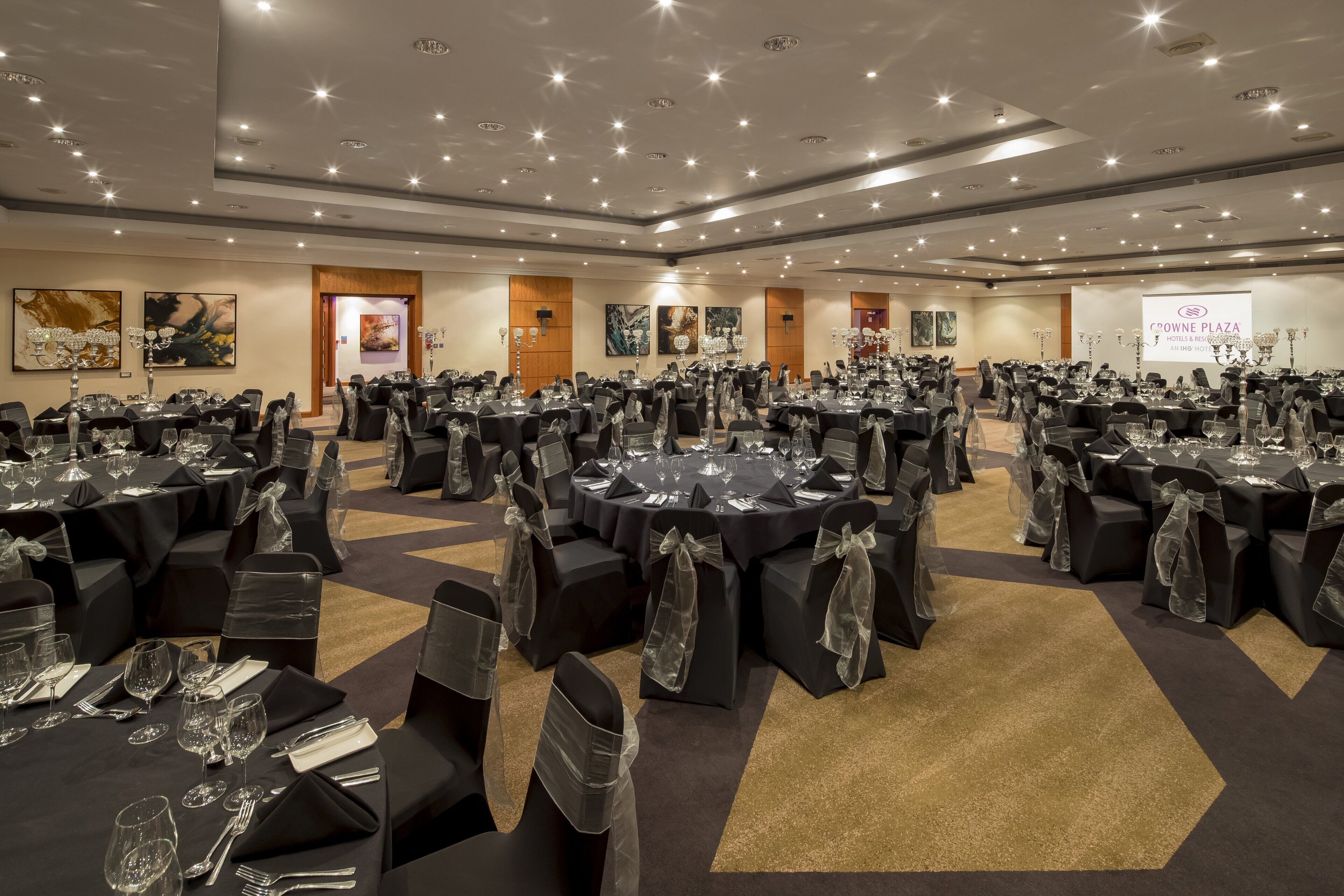 A large conference room with round tables, black chairs with clear sashes, and a carpeted floor.