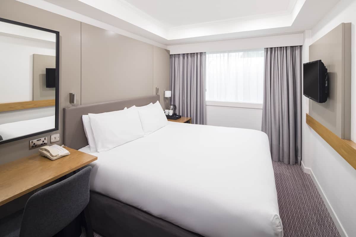 A hotel room with a large bed, a desk with a telephone, a TV mounted on the wall, and a window with curtains.