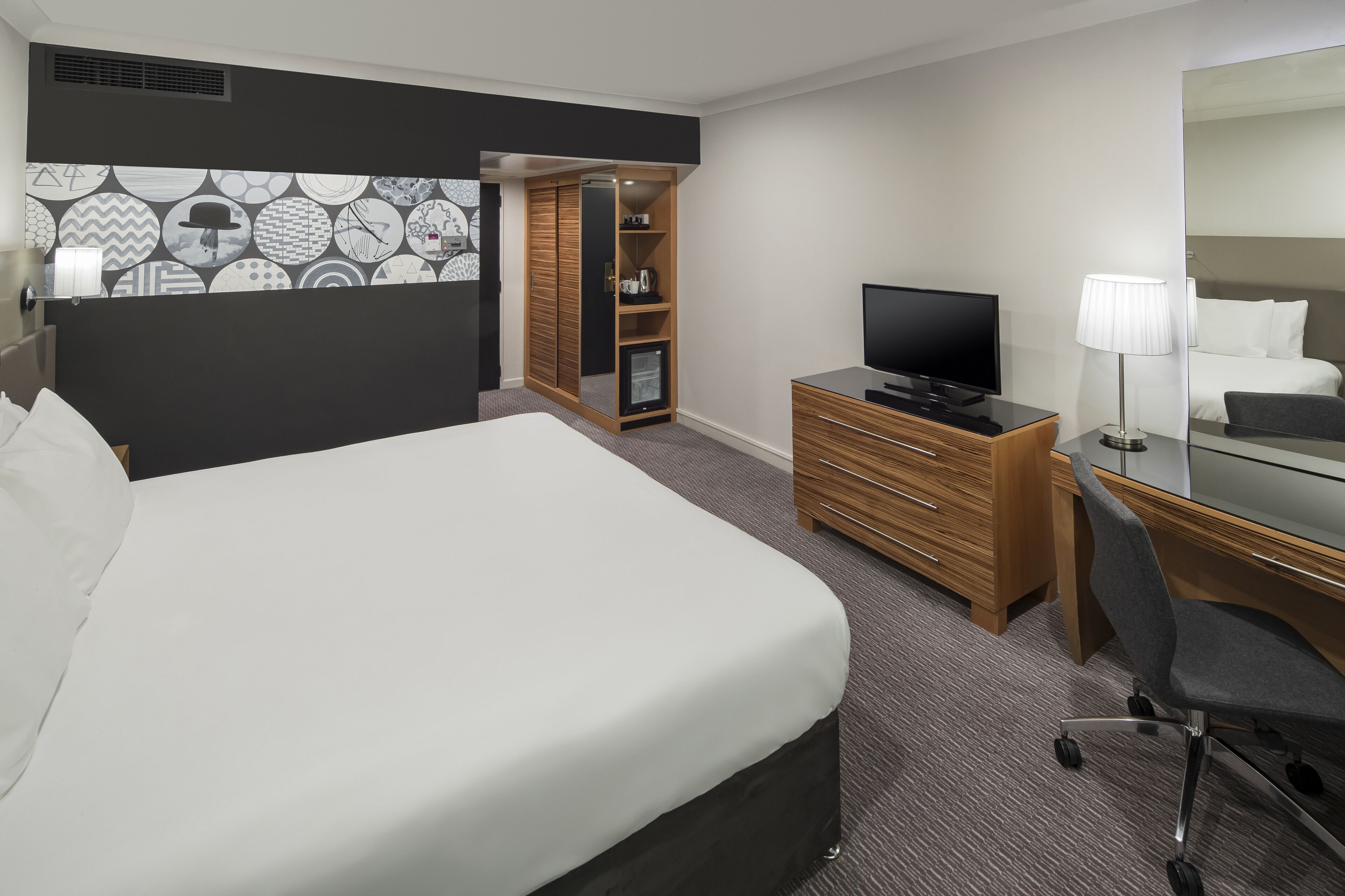Habitación de hotel con una cama grande, televisión, escritorio con silla, armario y espejo.