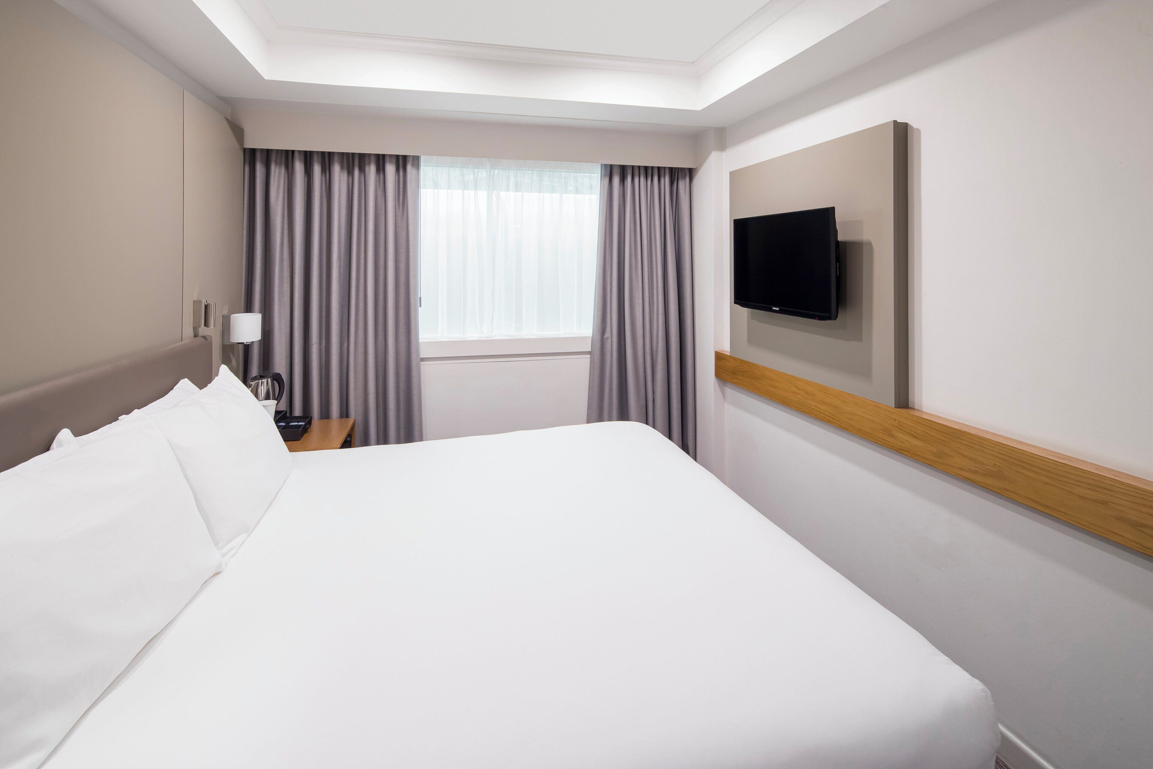 A hotel room with a large bed, a flat-screen TV mounted on the wall, a bedside table with a lamp, and a window with curtains.
