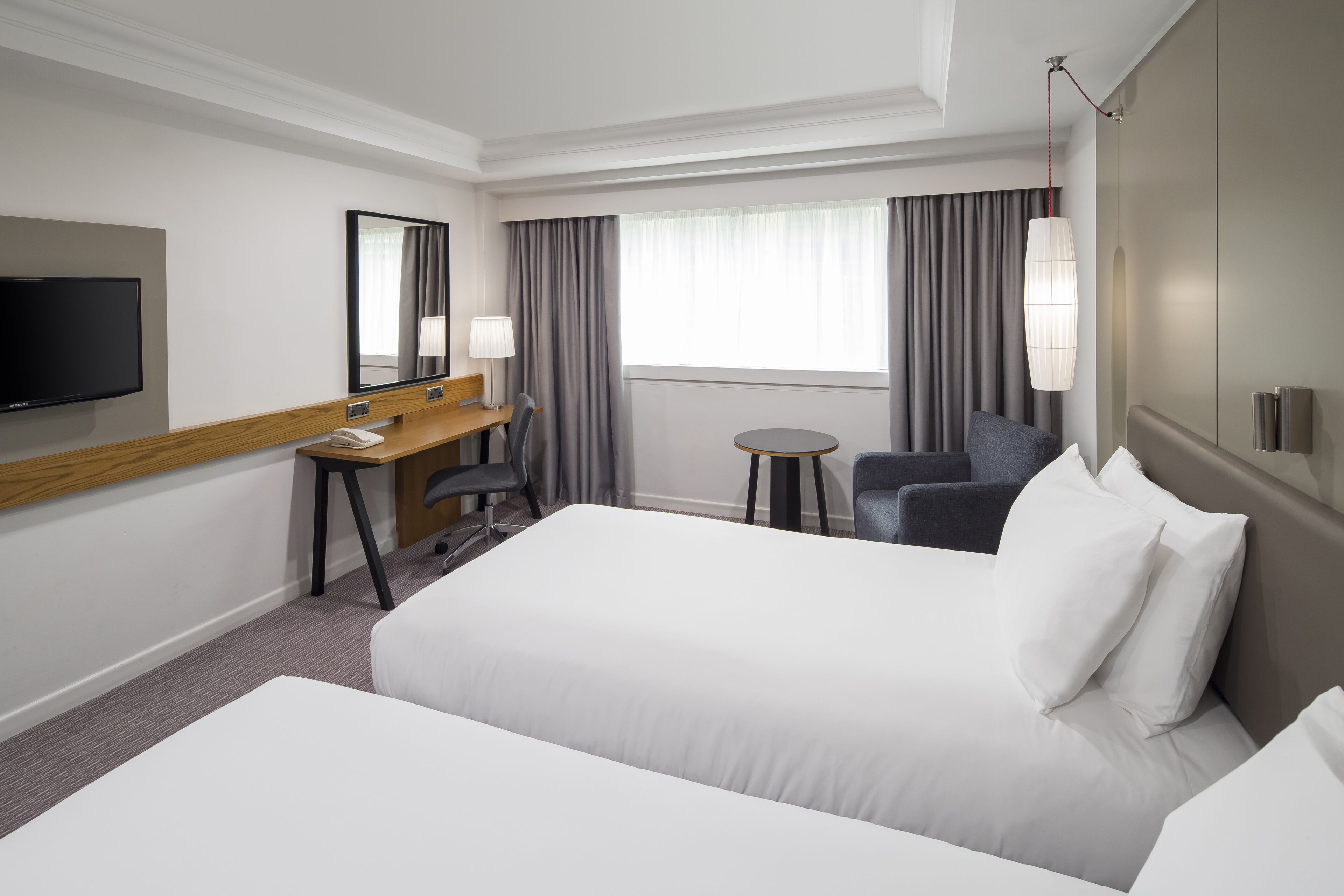 A hotel room with a bed, a desk with a chair, a TV, a small table, and a window with curtains.