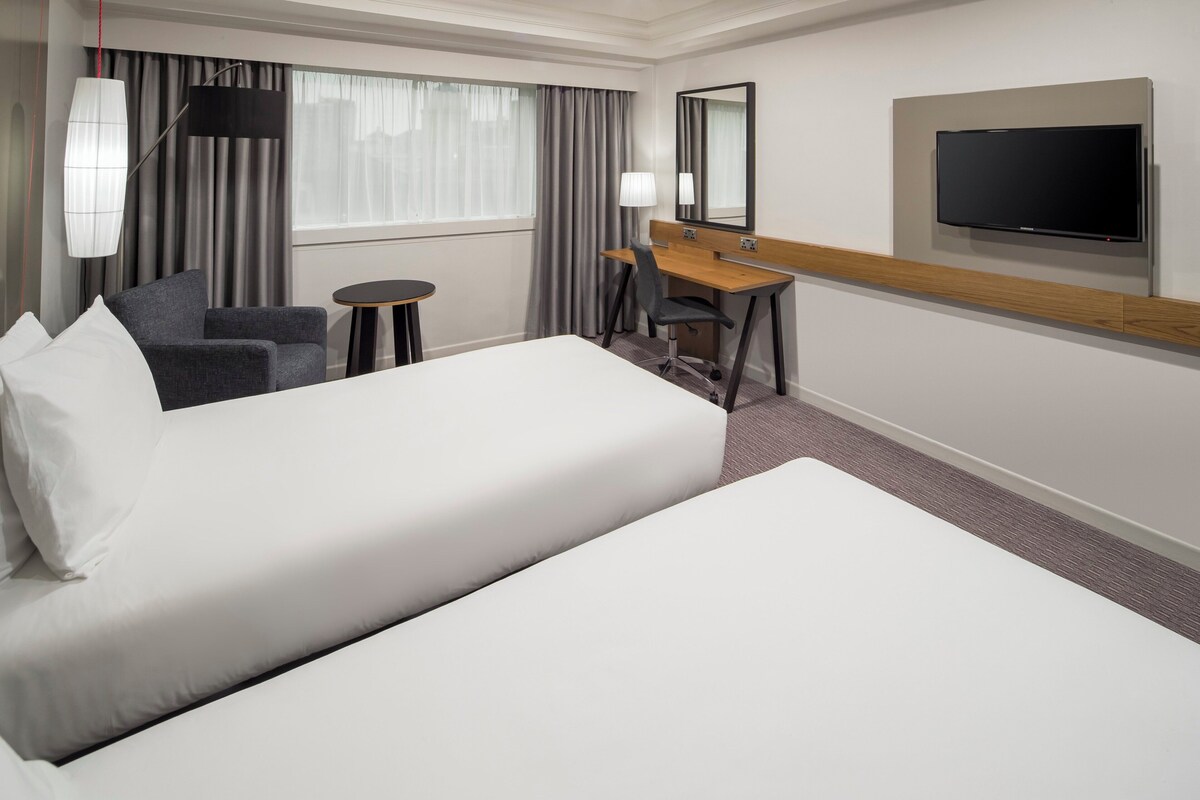 Ein Hotelzimmer mit zwei Betten, einem Schreibtisch, einem Stuhl, einem Fernseher und einem Fenster mit Vorhängen.