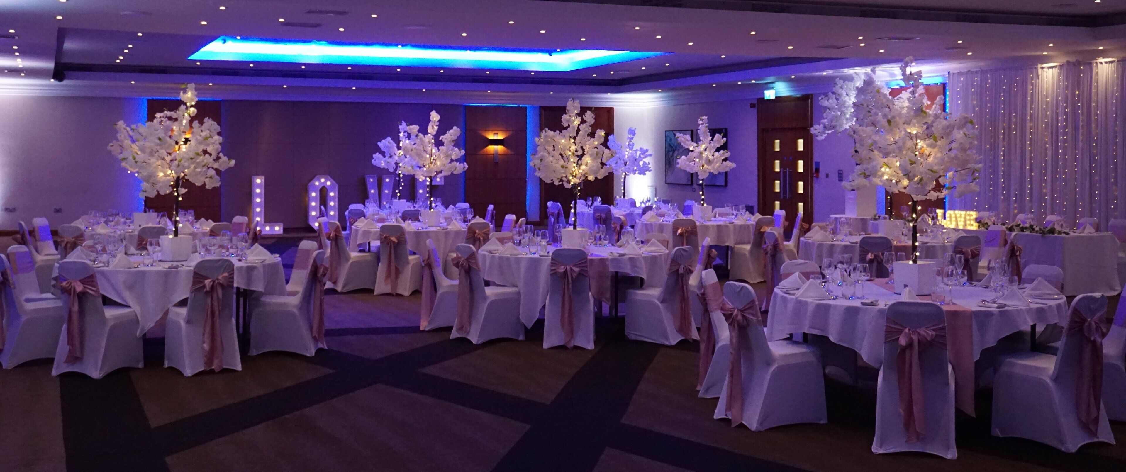 A banquet hall with round tables covered in white tablecloths, each with chairs tied with pink sashes.