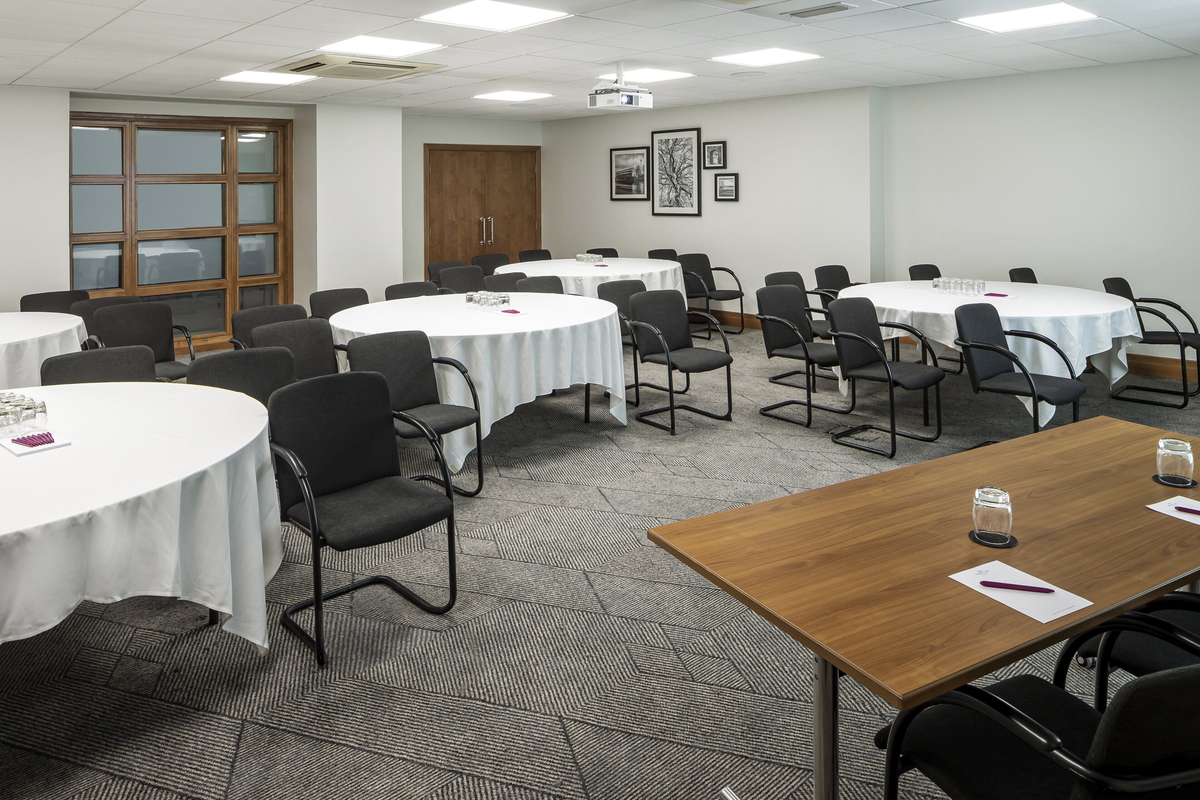 A conference room with multiple round tables, each with a white tablecloth and black chairs. There are framed pictures on the wall, a wooden door, and a carpeted floor.
