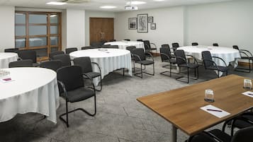 A conference room with multiple round tables, each with a white tablecloth and black chairs. There are framed pictures on the wall, a wooden door, and a carpeted floor.