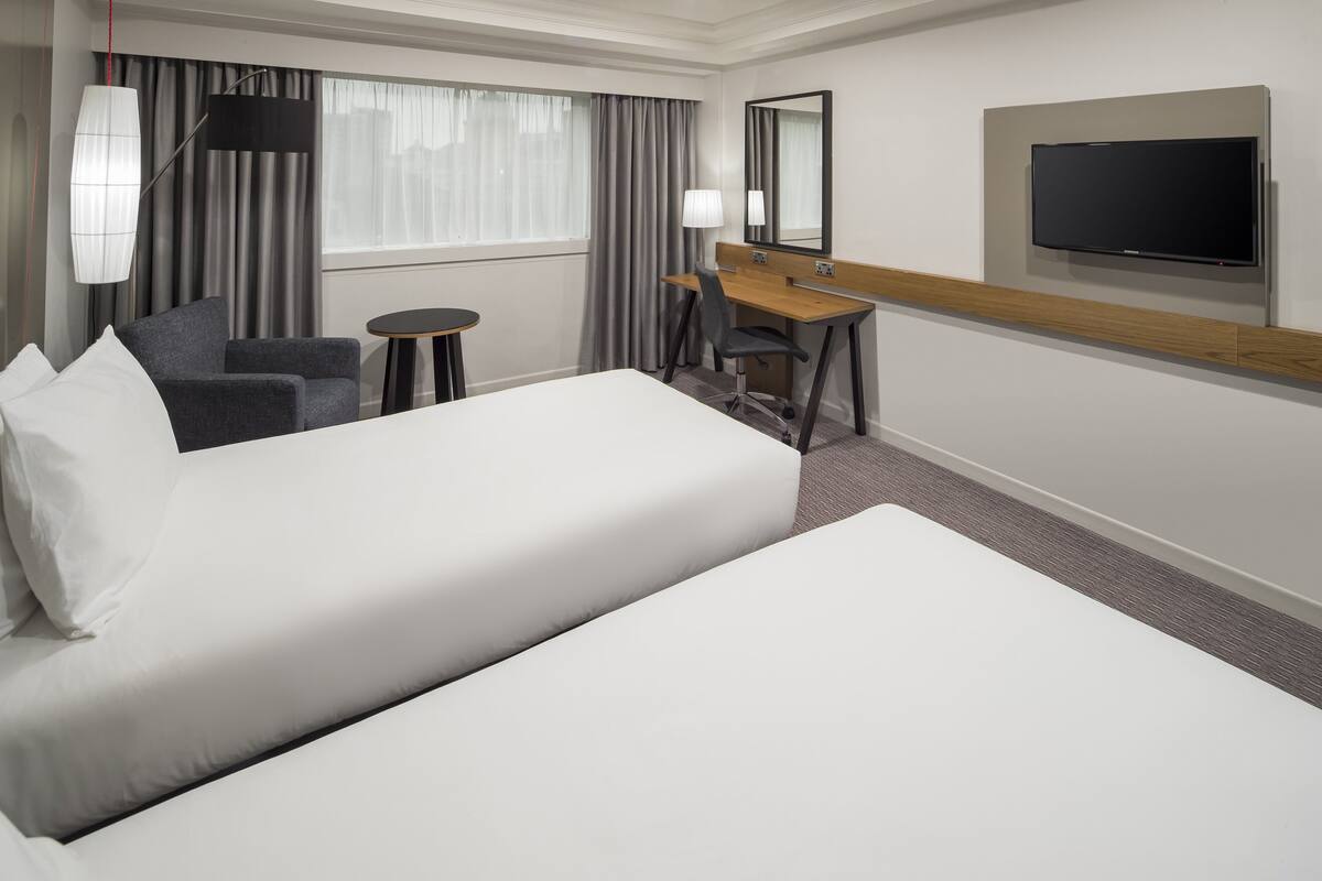 A hotel room with two beds, a desk, a chair, a TV, and a lamp.