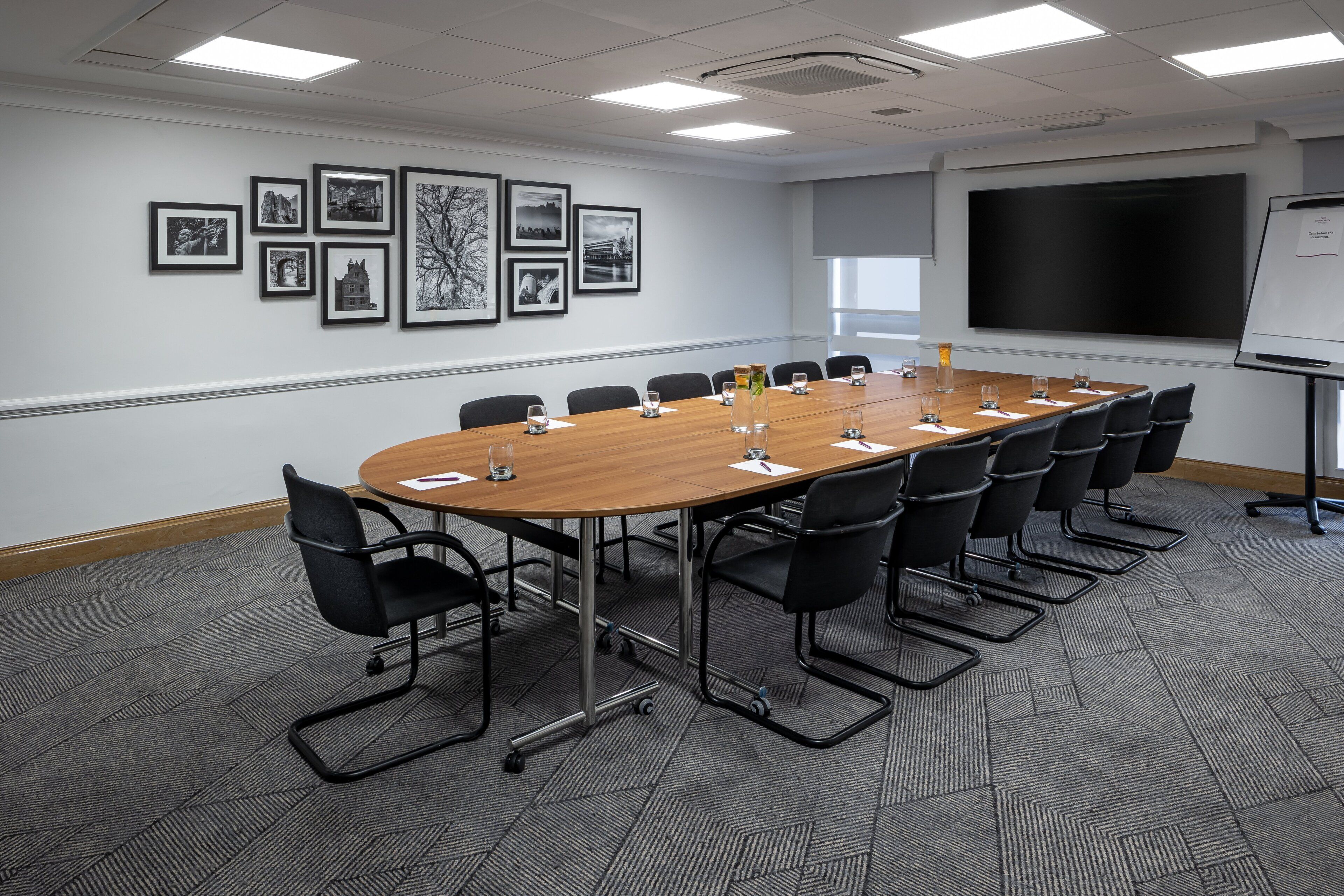 Ein Konferenzraum mit langem Tisch, schwarzen Stühlen, gerahmten Bildern an der Wand, einer Projektionsleinwand und einer Whiteboard-Tafel.